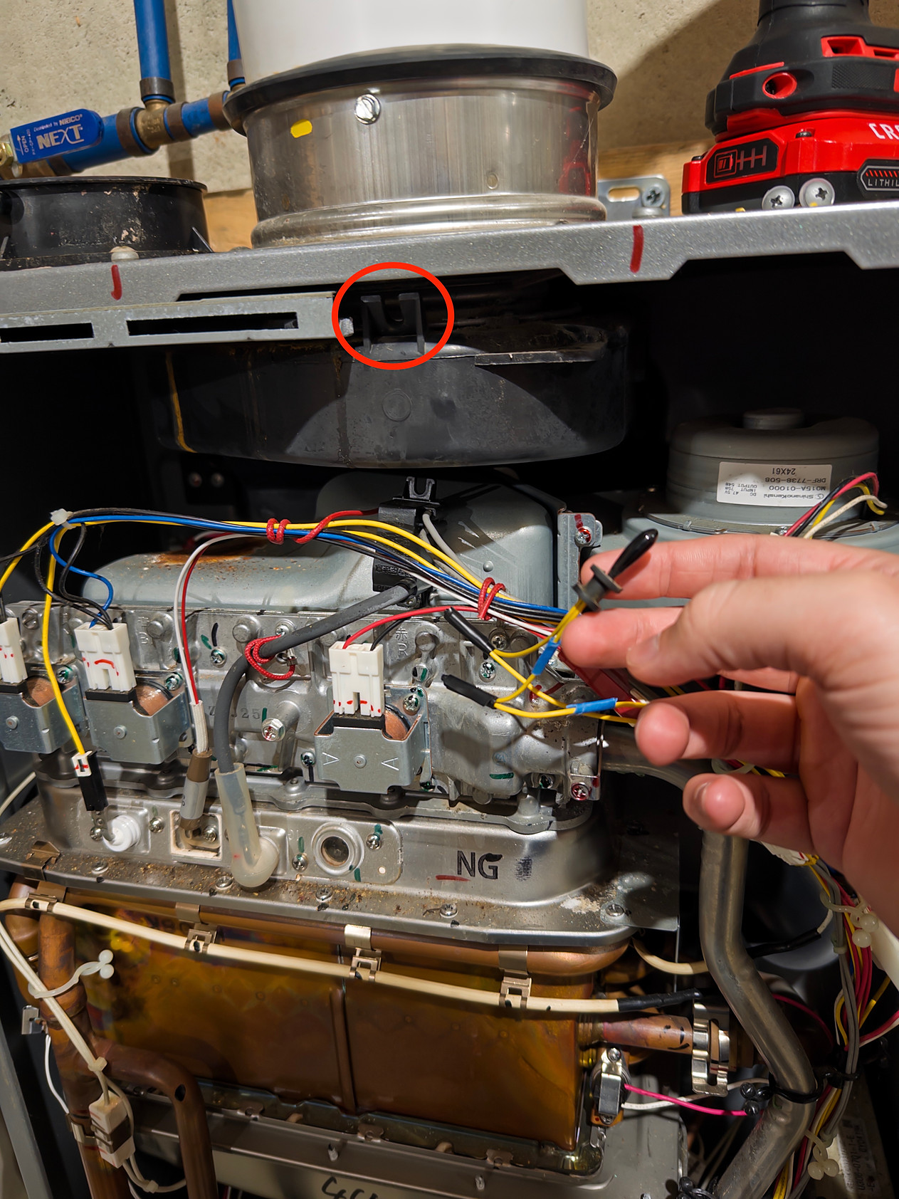 Top of the water heater's interior below the exhaust collar, with the sensor's mounting clip circled in red, and a hand holding the small black sensor with its yellow wires spliced using blue butt connectors